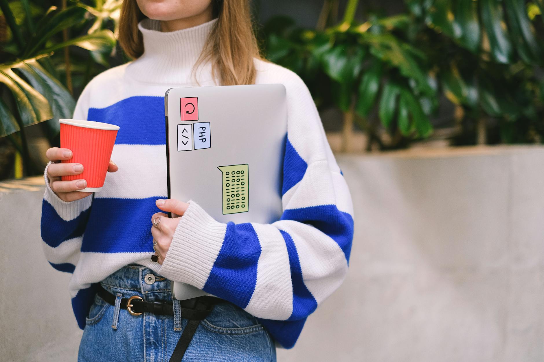 Woman in striped sweater holding laptop and coffee cup, exuding casual student life.
