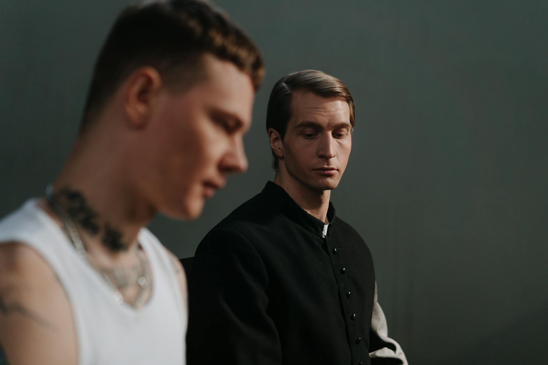 Two men in conversation, one in a priest's attire, indoors with a contemplative mood.