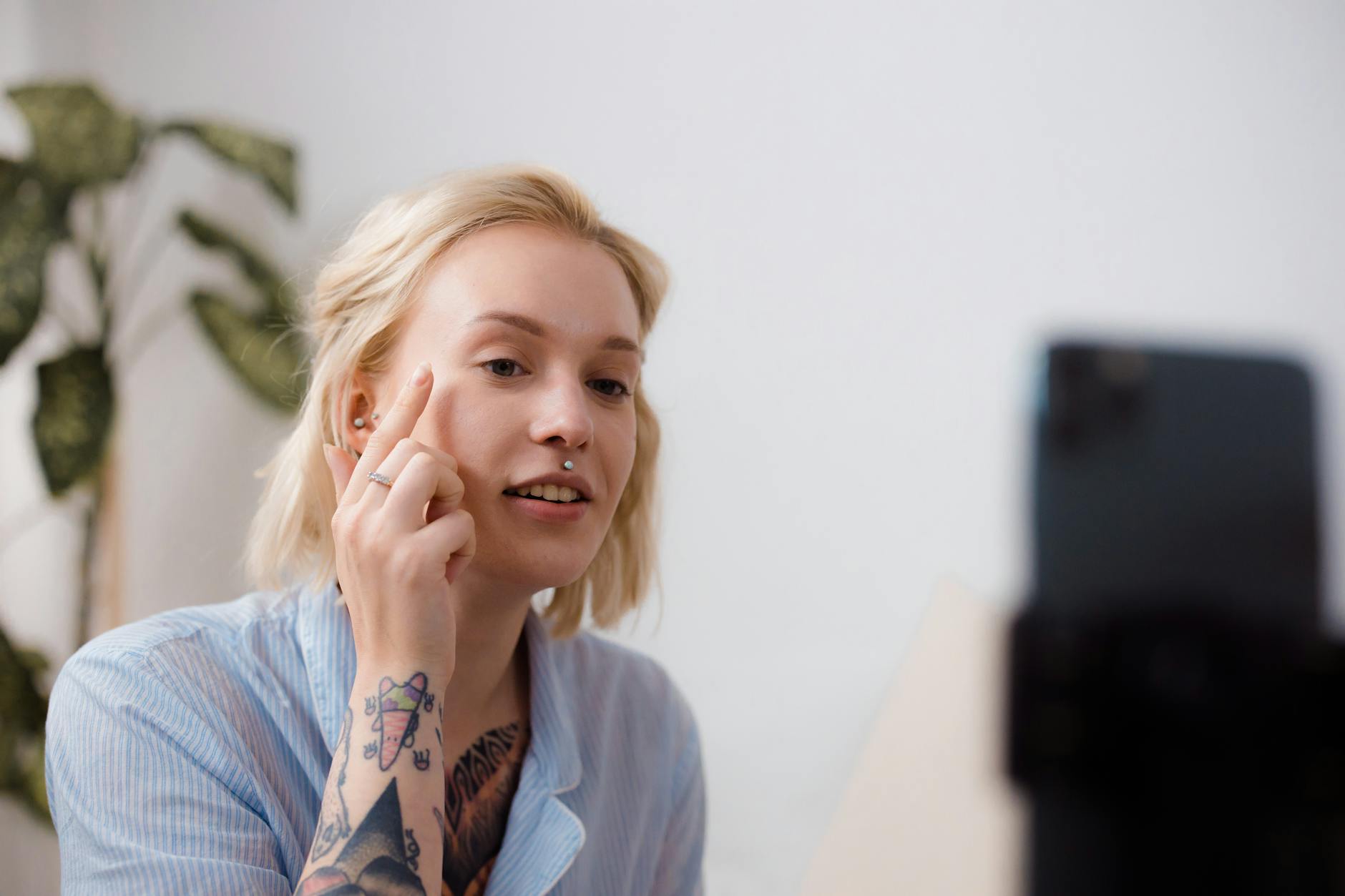 A young woman with tattoos recording a makeup tutorial with a smartphone indoors.