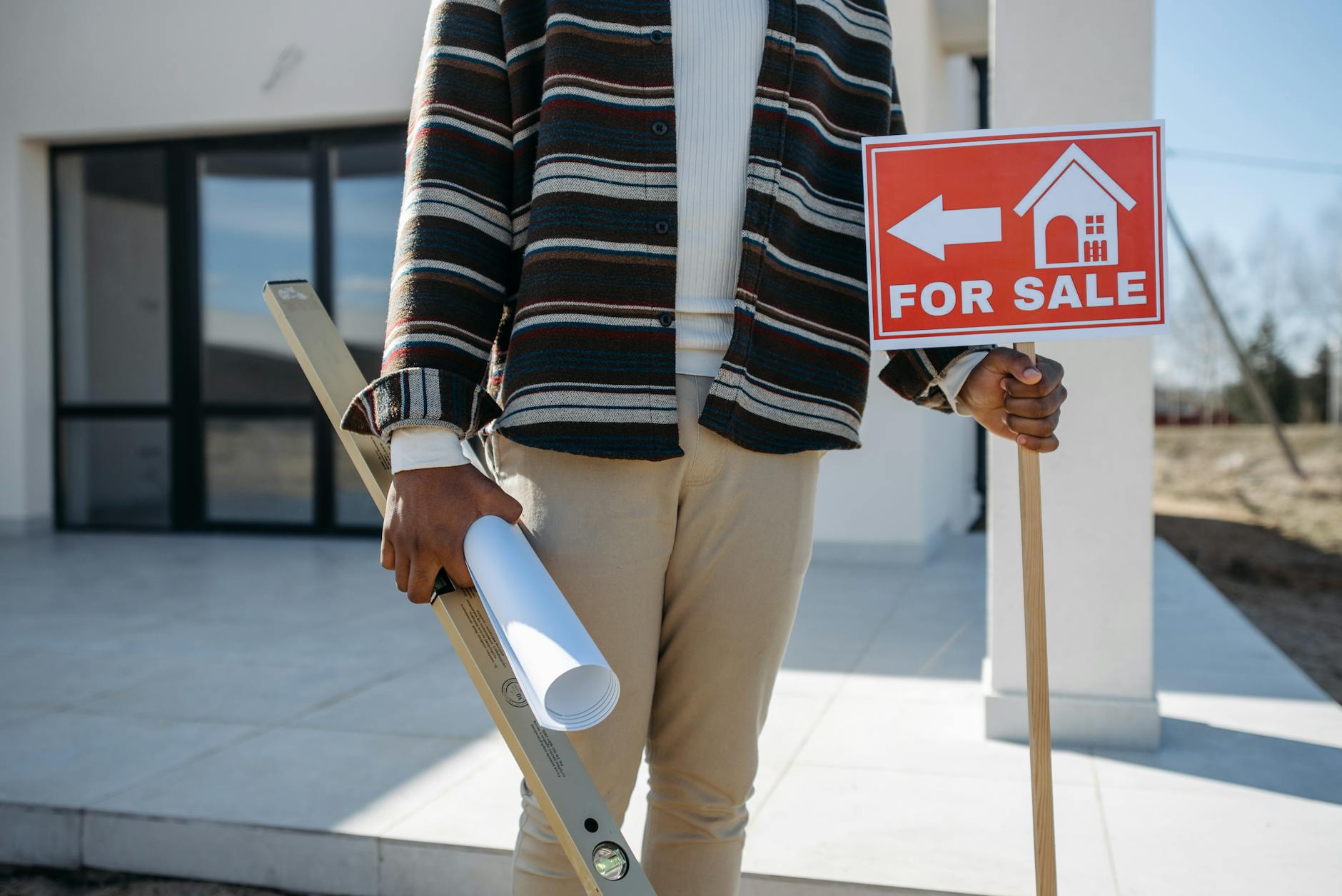 A real estate agent stands outside with a 'for sale' sign and rolled blueprint, suggesting a property for sale.