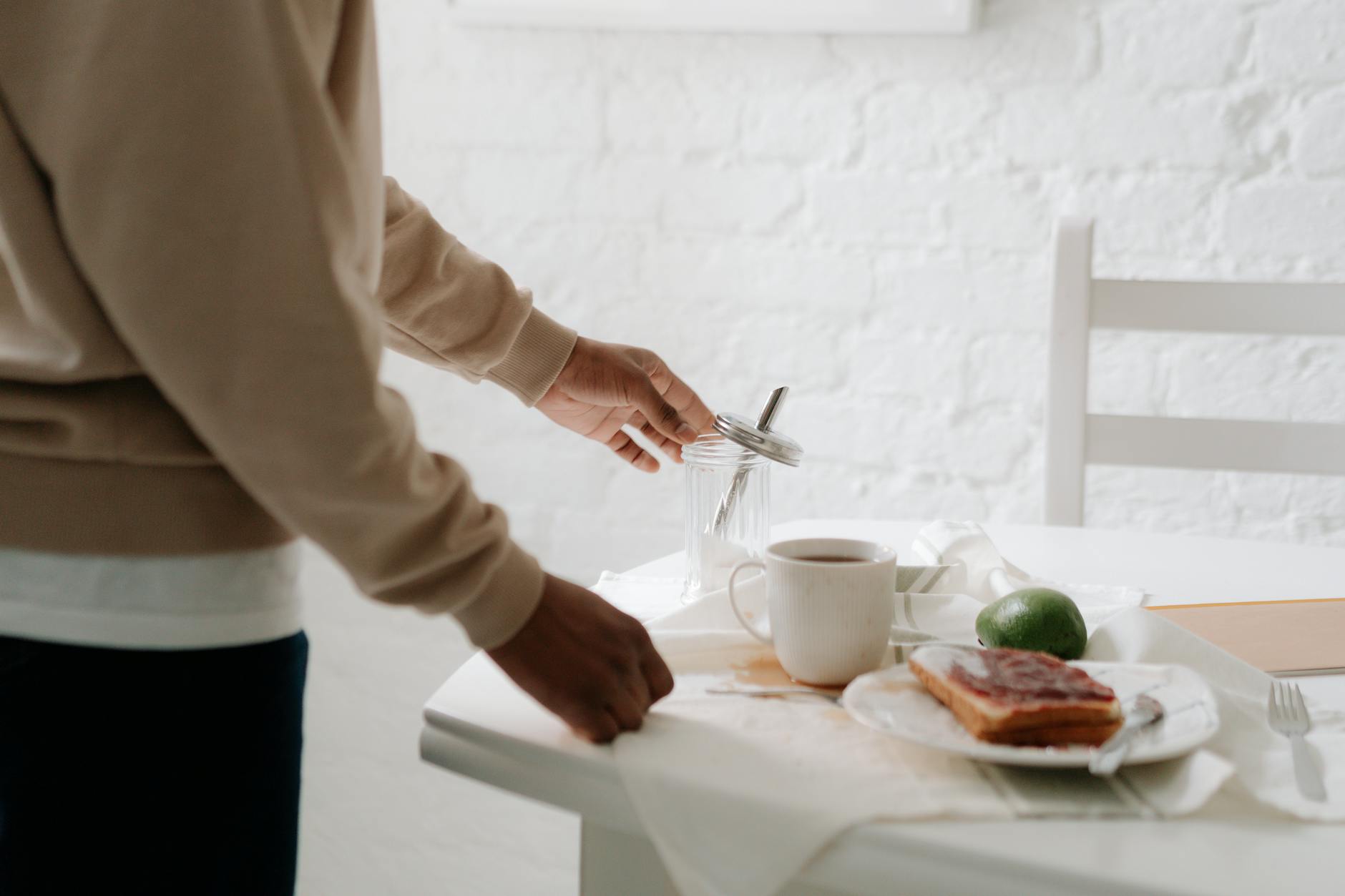 A person preparing a breakfast table with coffee, jam toast, and avocado indoors.