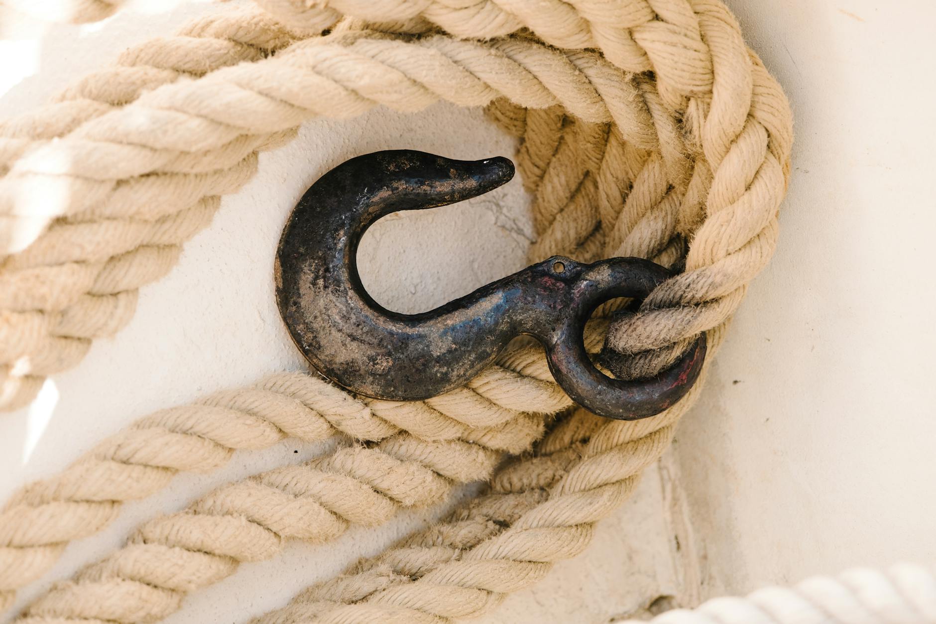 Close-up of a rustic metal hook entwined with coiled beige nautical rope on a white surface.