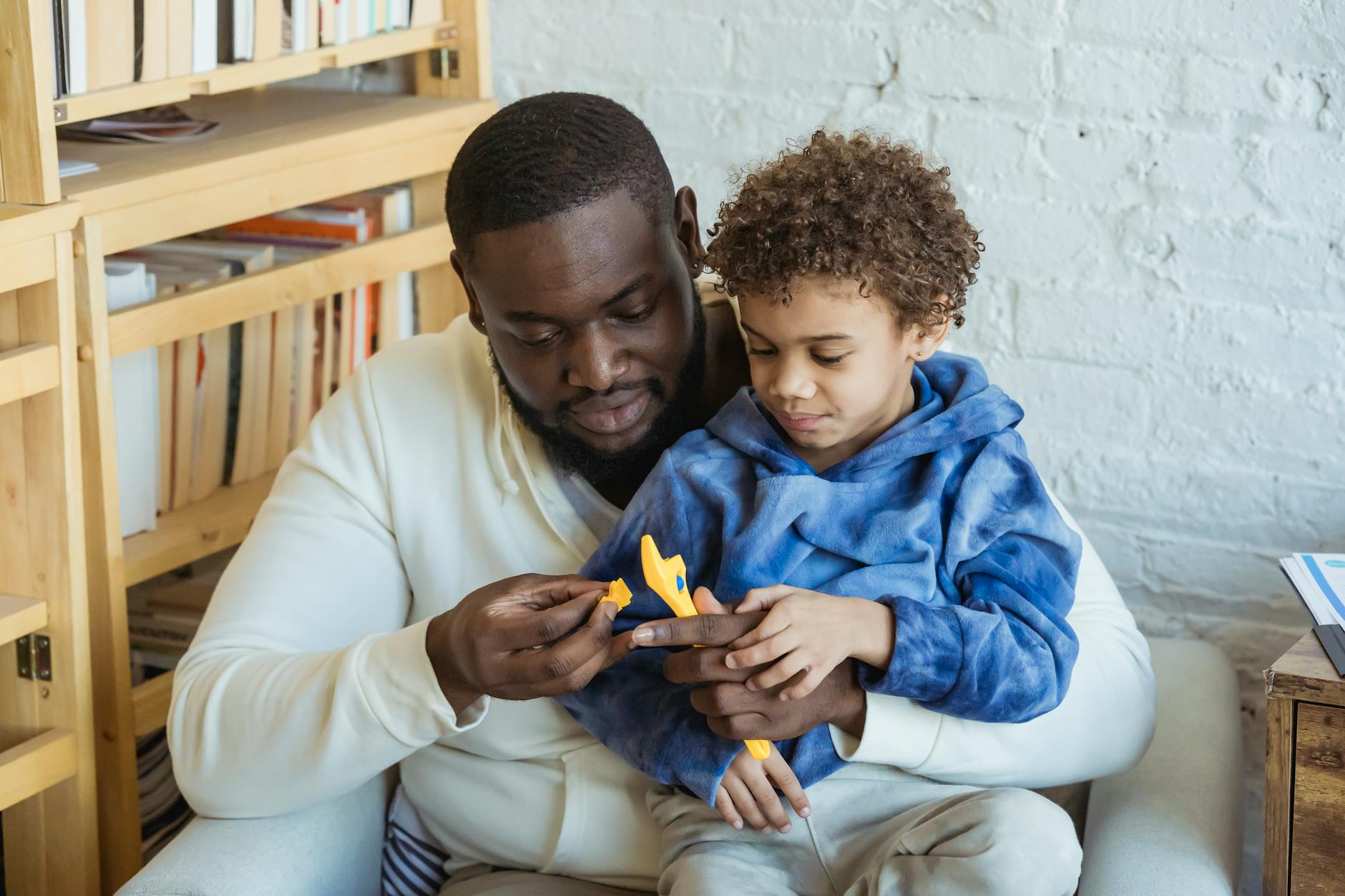 Curious African American father and black son with toy wrench sitting near shelves while playing in light room at home