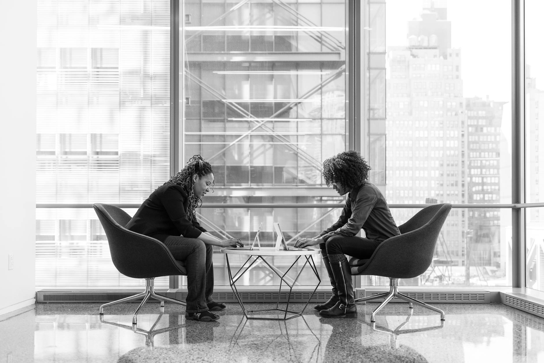 Two women engaged in a business discussion at a modern office, sitting across a table with a city view.